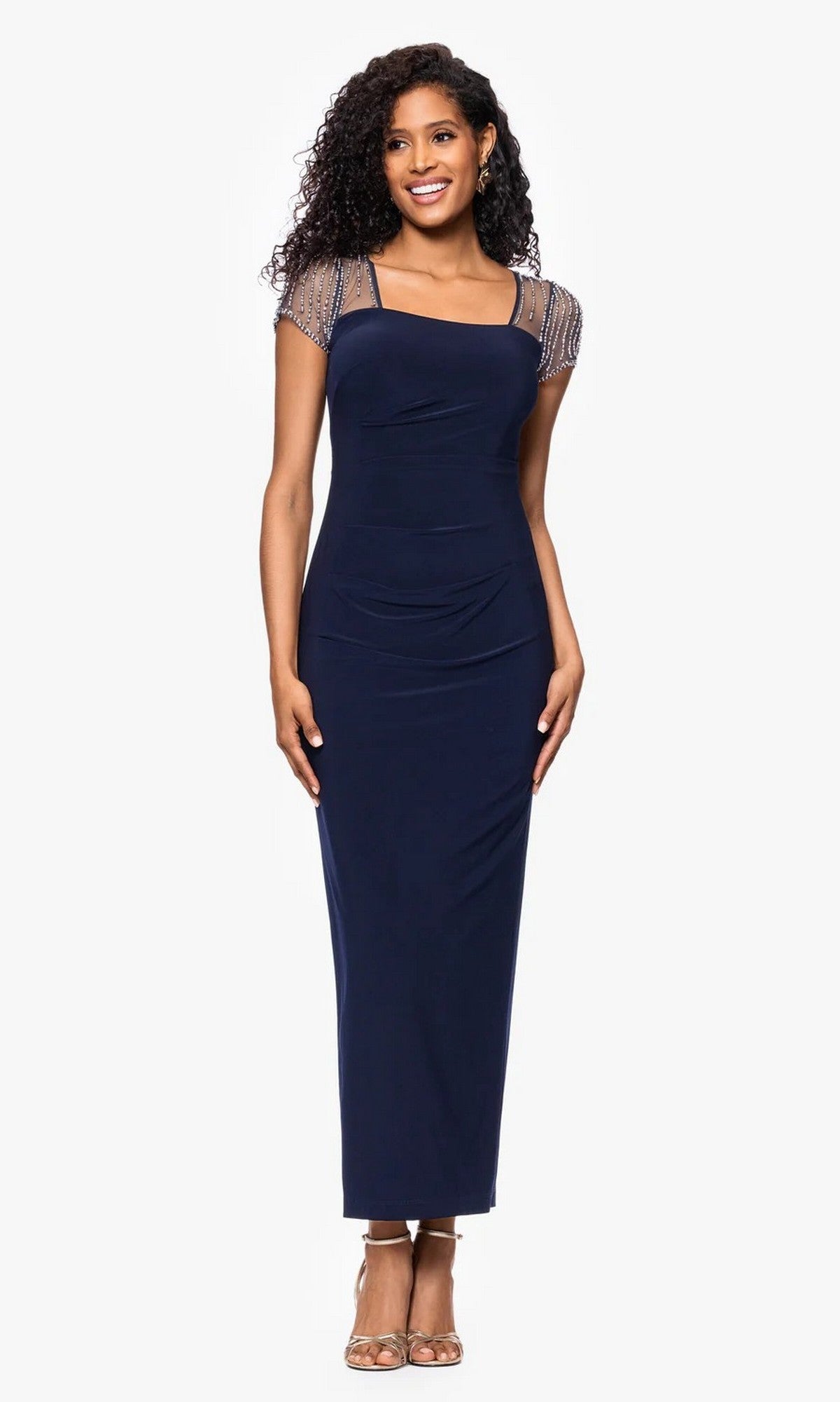 A woman wears the Long Navy Formal Dress: Betsy & Adam A25994 in navy/silver, featuring beaded sheer short sleeves and beige heels, standing against a plain white background.
